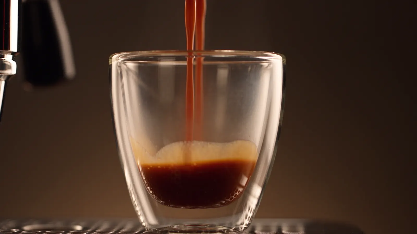 Un espresso coule dans un verre à double paroi, sur un fond brun sombre.